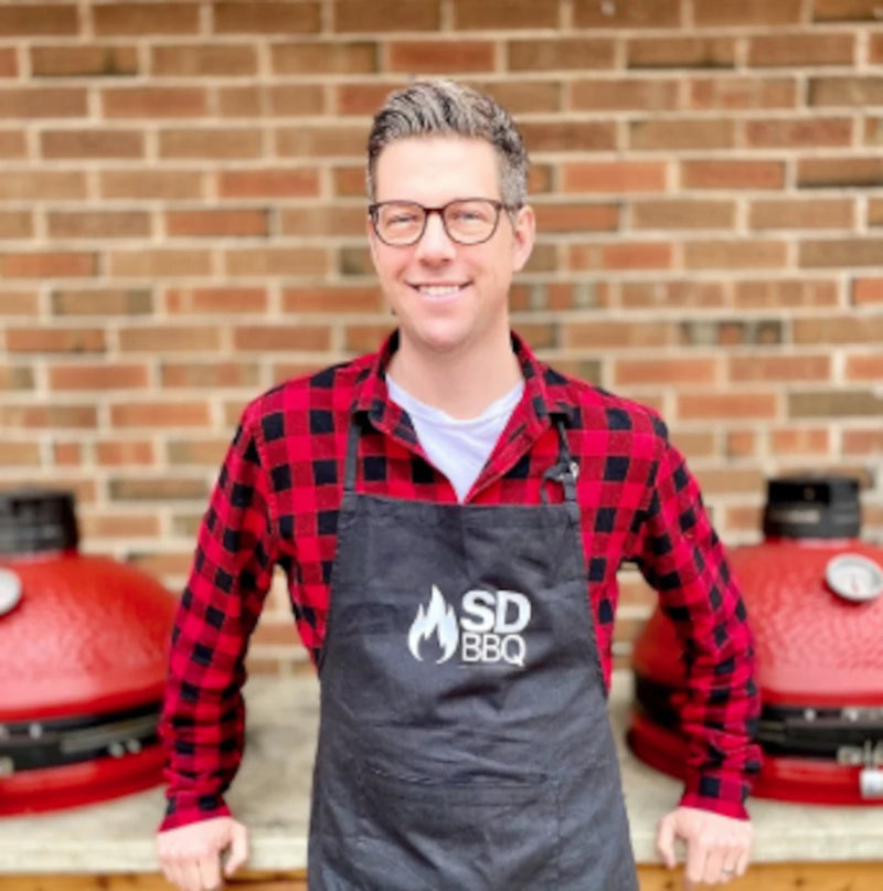 James Mack wearing a Smoking Dad BBQ apron over a red and black checked shirt leans against a counter with 2 built-in Kamado Joe grills installed in it.