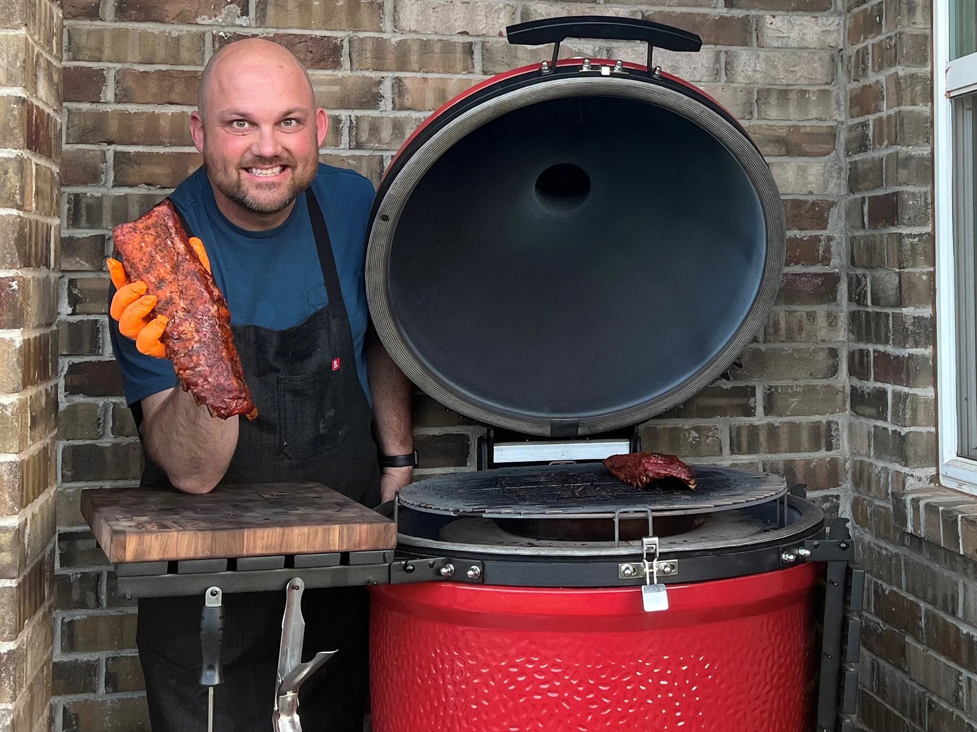 Scott Bartlow smiles while standing next to an open Kamado Joe grill. Scott is holding up a cooked rack of ribs.