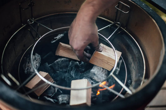 Someone reaches down into a Kamado Joe grill to stack wood chunks on top of freshly lit charcoal