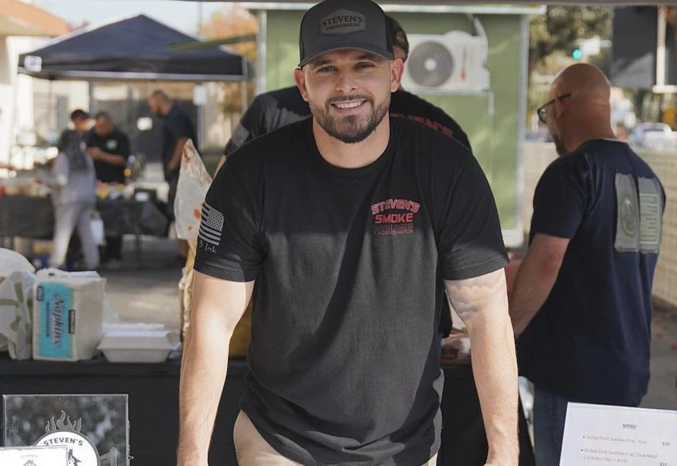 Steven Odum at an outdoor BBQ festival wearing a black t-shirt and cap, each with the Steven's Smoke House logo.