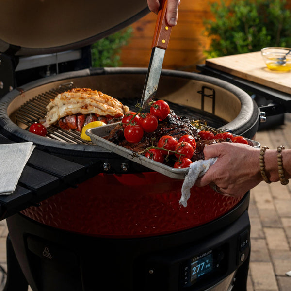 Someone uses tongs to remove grilled tomatoes on the vine, steak, and lobster from the Divide & Conquer cooking racks in a Big Joe Konnected Joe grill