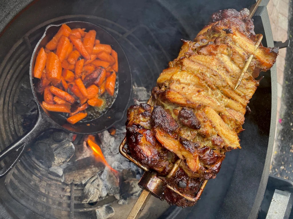 Chicken shawarma roasts on a JoeTisserie while a cast iron skillet of sauced carrots cooks on the coals to the side